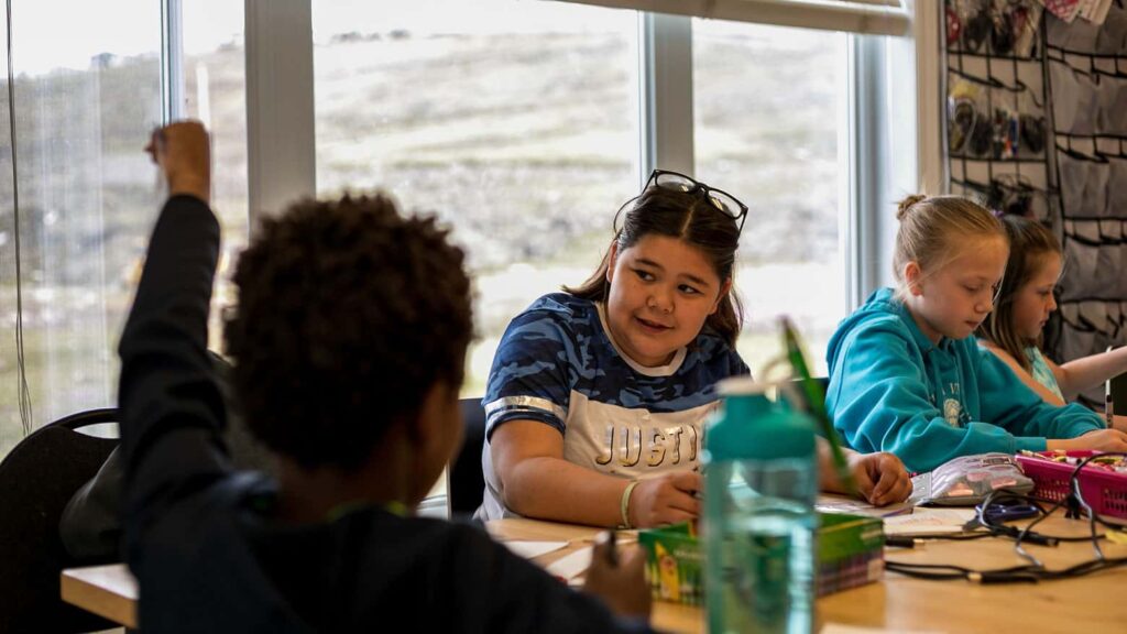 Kids looking at each other while sitting at a table with a big window behind them. One is facing the camera